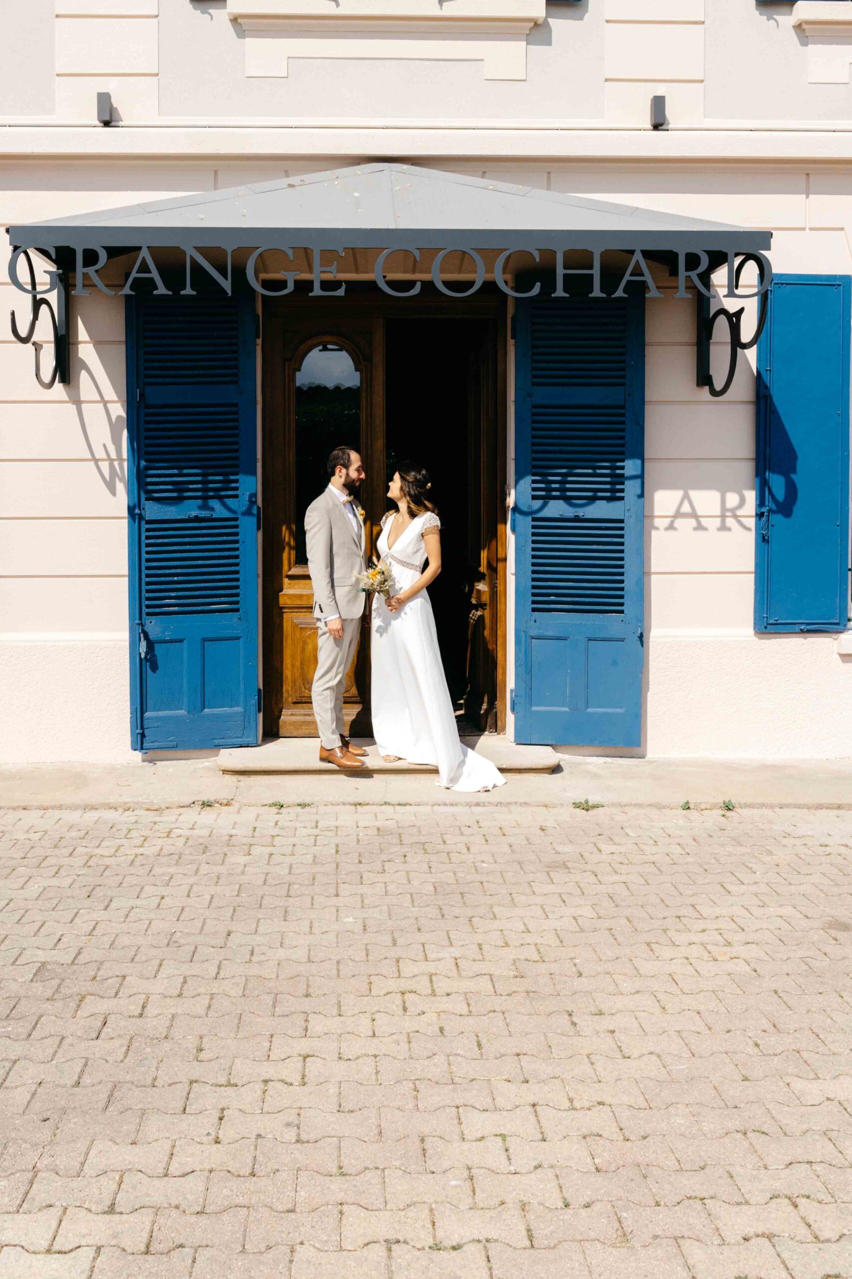 les mariés posent l'un face à l'autre devant l'entrée du château aux volets bleus, et dont les lettres provoquent des ombres sur la façade