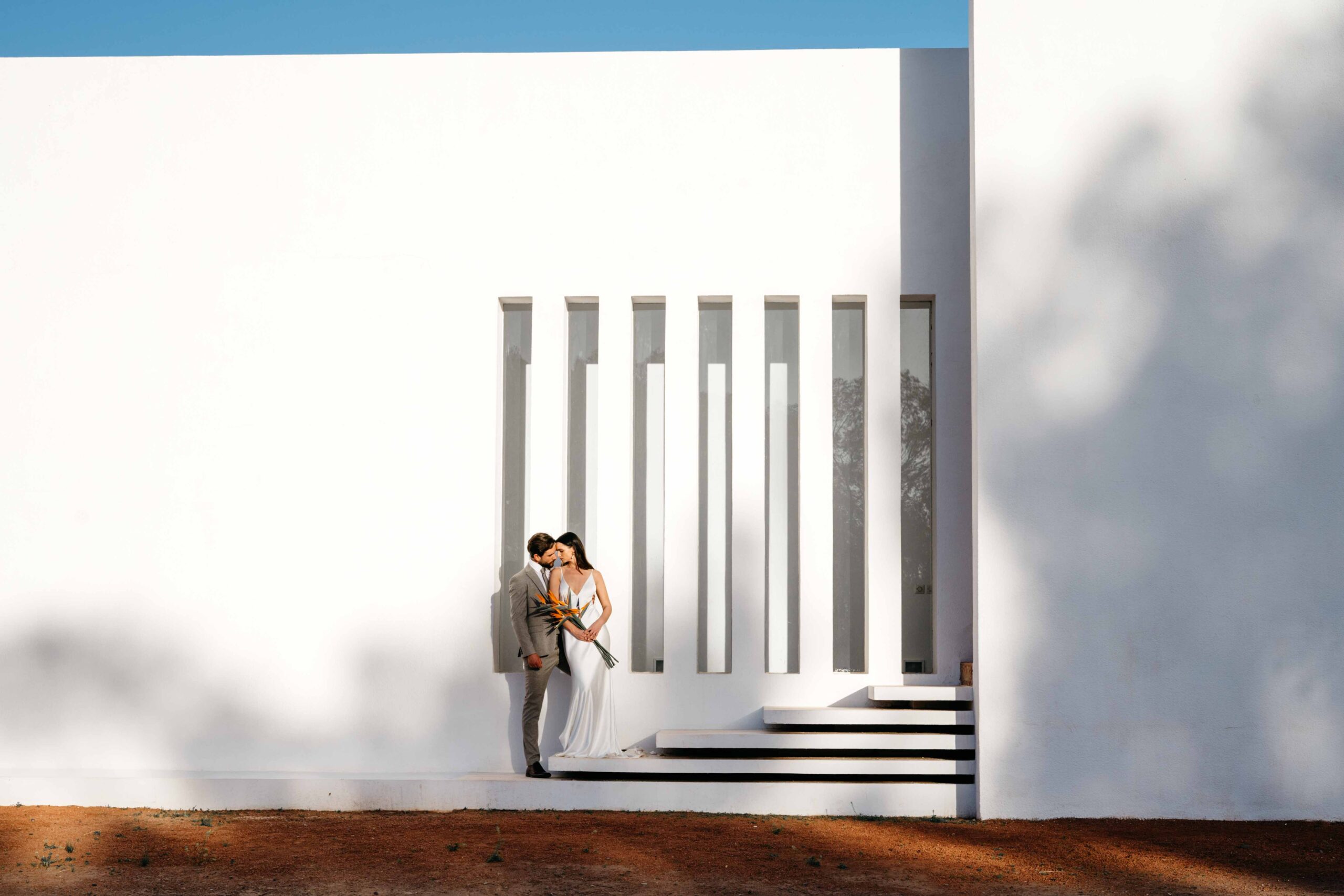 mariés s'embrassant devant la façade d'une maison architecturale moderne blanche. le plan photo est large pour ajouter une bande bleue du ciel en haut, et une bande oranger du sol en bas.